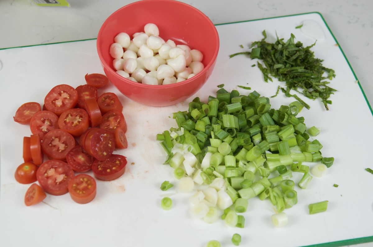 Diced tomatoes, mozarella, chopped green onion, chopped mint and chopped basil on a white cutting board
