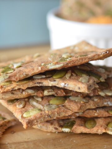 Stack of homemade seed crackers on a wooden board with a dip cup in the background