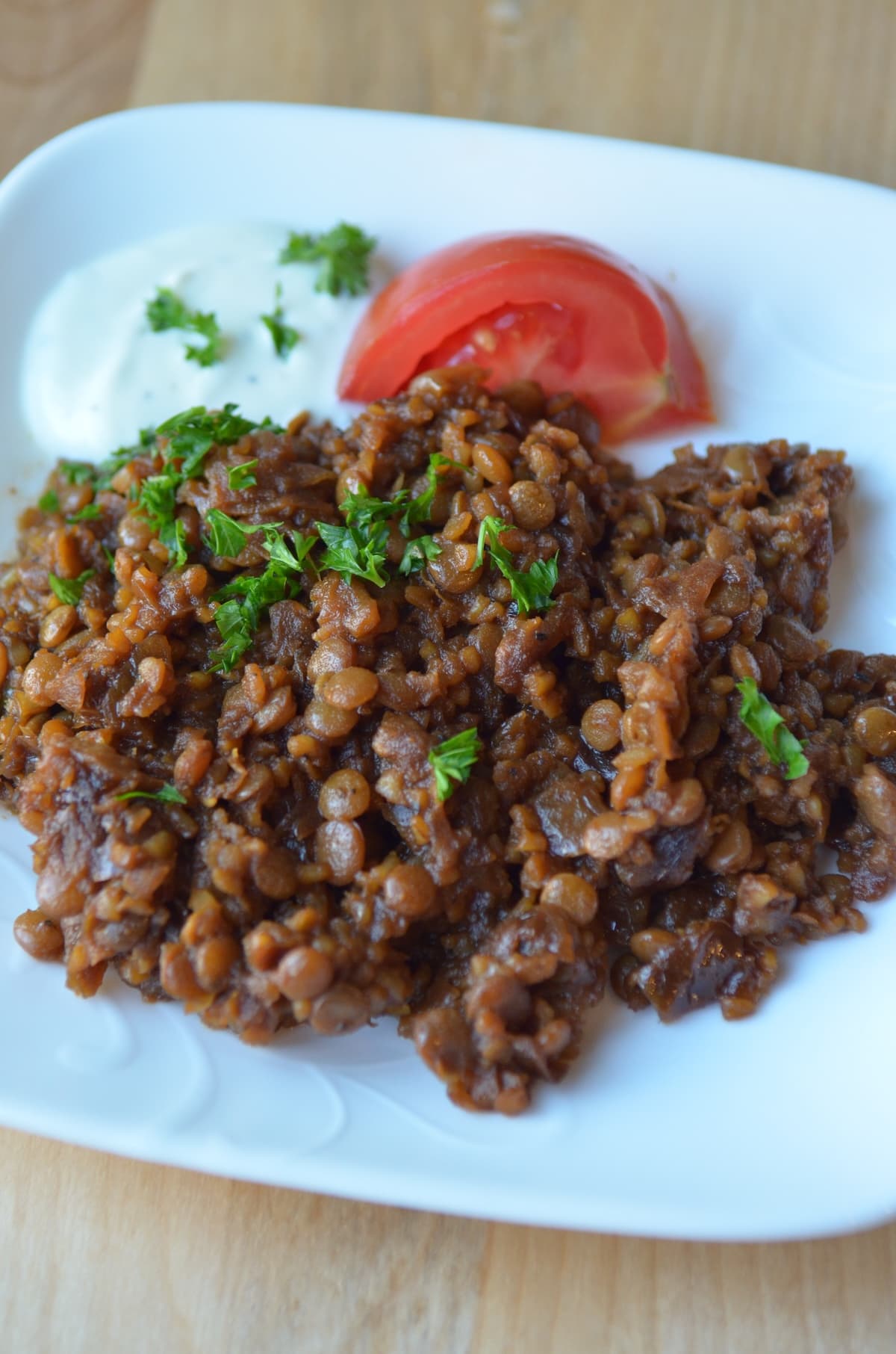 A closeup shot of mujadra hamra on a white plate, sprinkled with parsley and garnished with yogurt sauce and tomato.