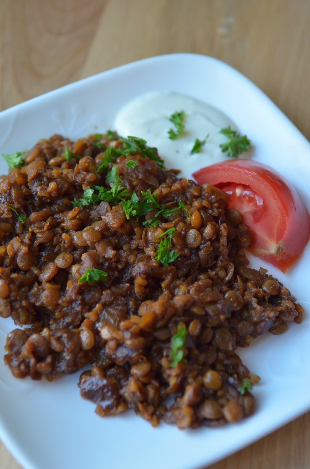 A square plate of mujadra hamra, sprinkled with parsley and garnished with yogurt sauce and tomato.